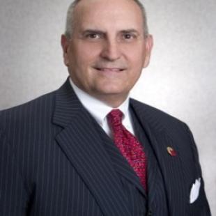 Man with thin gray hair in black pin-striped suit with red tie.
